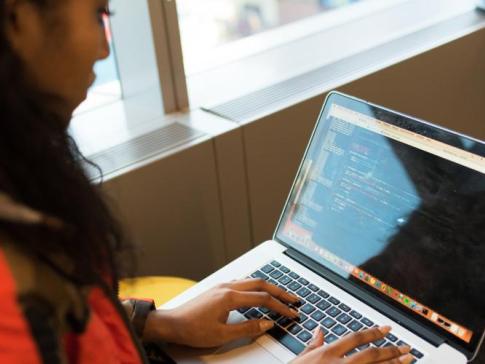 A photograph of a young women of color sitting in a chair and typing on a laptop