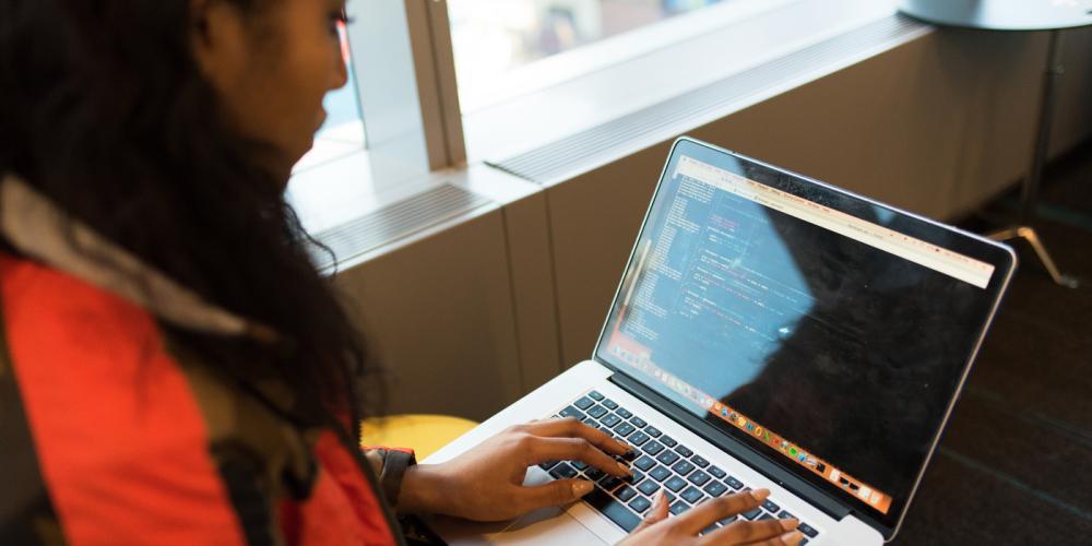 A photograph of a young women of color sitting in a chair and typing on a laptop